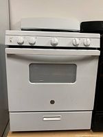 Front view of the GE white gas stove showing door with window, control knobs, and bottom drawer.