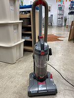 Full view of the upright Hoover vacuum showing the handle, flexible hose, and base in a grey and red color scheme.