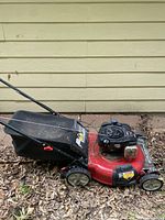 Side view of red Murray lawnmower with attached black grass catcher bag on a bed of dry leaves near wall siding, showing the handle, wheels, and engine assembly.