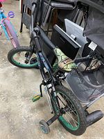 Full angled view of the black kids bike showing the TMNT-themed seat, green rims, black tires with training wheels attached.