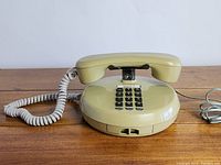 Front and side view of olive green push button telephone with coiled handset cord and straight line cord on wood surface.