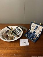 Photo showing the Swinnertons transferware bowl filled with various jewellery items and small decorative objects, next to a closed vintage blue fabric-lined case holding silver plated spoons and a small plated dresser box.