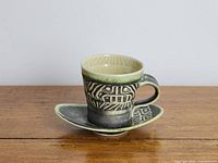 Green pottery cup and saucer with First Nations style decorative patterns, shown on wooden surface against plain background.