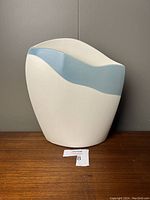 Front view of the signed John Bergen ceramic vase showing its white body and curved blue band design, placed on a wood surface against a gray wall.