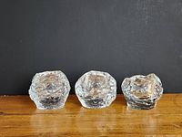 Front view of 3 clear glass snowball style candleholders on wooden surface with black background.