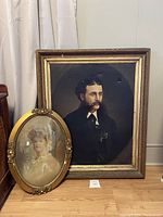 Full view of the two portraits placed against a wall showing the large oil painting and the oval framed photograph