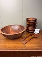 Wooden large salad bowl, nine smaller bowls stacked, and two wooden salad tossers on a wooden surface against neutral background.