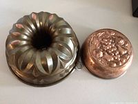Two bundt pans on a white surface: larger aluminum bundt pan on left with smooth fluted swirl design, smaller copper bundt pan on right featuring embossed fruit pattern.