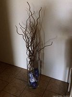 Front and side view of a clear cylindrical glass vase about 20 inches tall, with multiple blue and white patterned bulbs inside and tall brown twisted decorative branches extending upward.