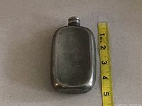 Top down view of small rectangular silver flask next to measuring tape showing about 4 inch length