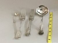 Small salt spoon, two forks (one larger with squared end, one smaller with shell motif), and a gravy ladle with detailed handle design arranged with a ruler for scale.