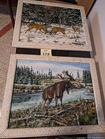 Photo showing two framed wildlife oil paintings: one of deer in snow and one of a moose by a river.