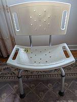 White adjustable assisted bath chair with perforated seat and back, aluminum legs, and black rubber feet placed on a carpet near a curtain.