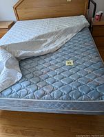 Photo showing queen size mattress partially covered by a white mattress protector, wooden headboard visible at the top, matching box spring under mattress, hardwood floor visible.