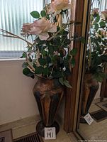 Photo showing a tall brass urn vase with a bouquet of artificial pink roses and leaves placed inside, next to a mirror and window.