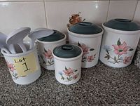 Photo showing five ceramic cannisters of varying sizes with floral designs and green lids, plus a ceramic holder containing assorted white plastic cooking utensils.