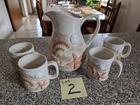Ceramic pitcher and four matching mugs on a stone counter, showing embossed seashell and starfish designs on each item.