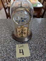Front view of 9-inch cloche battery operated clock under glass dome, showing clock face with gold rim, multiple dials, blue hands, and gold pendulum balls inside.