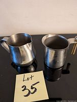 Photo of a stainless steel creamer with handle and spout next to a taller stainless steel sugar bowl without a lid