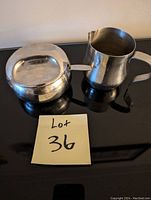 Photo of two stainless steel serving pieces: a creamer with spout and handle, and a lidded sugar bowl with handle, placed on a black reflective surface next to a note with 'Lot 36'.