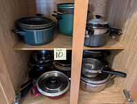 Wooden cabinet with two shelves filled with assorted pots and pans including enamel, metal, and frying pans with lids.