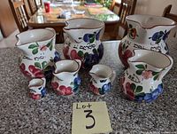 Six decorative ceramic pitchers arranged in a growing size order from smallest (about 2 inches) to largest (about 6 inches) on a granite countertop. Each pitcher features colorful floral artwork and Italian writing on the front with red rims.