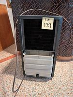 Photo of a portable dehumidifier unit showing the water collection tank and a power cable on a terrazzo floor.