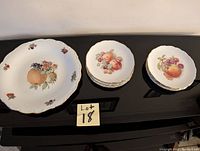 Six gold rimmed white dessert plates and one matching serving platter, all decorated with detailed fruit illustrations, scalloped edges, and gold rims.
