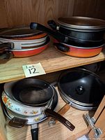 Photo of assorted frying pans of various sizes and styles on a wooden shelf, showing wear and scratches.