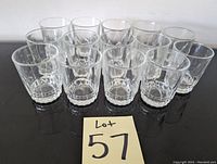 Photo showing 14 clear short drinking glasses arranged in two rows on a black reflective surface with a lot number card in front.