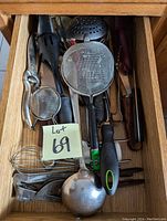 Top view of a drawer containing various metal and plastic kitchen utensils including strainers, ladle, whisks, and other tools.