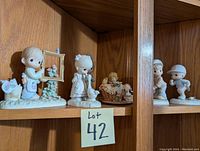 Wooden shelf with five assorted Precious Moments porcelain figurines positioned spaced apart.