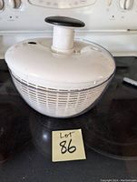 Photo of a white lid salad spinner on a black stovetop with the lid closed and a black handle on top. The clear bowl and interior basket are visible through the sides.