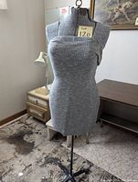 Gray fabric-covered sewing mannequin model or dress form with a tripod metal stand in a room, used for sewing and tailoring.