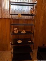 Front view of black metal shelving unit with six shelves placed against a wood paneled wall with a curtained window behind it. The shelves hold a few small decorative items.