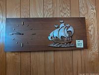 Photo of rectangular wooden wall clock with metal sailing ship decoration and minimalist clock face mounted on wood panel wall.