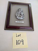Framed wall hanging showing Madonna holding baby Jesus, mounted on beige mat board within dark wood frame with gold trim. Includes a metal plaque with artist name.