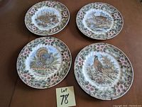 Four ceramic decorative wall plates laid out on a wood surface with a lot number card '78'. Each plate features a different wildlife scene (deer, turkey, pheasant, duck) surrounded by a pink floral border. The plates include hangers for wall display.