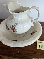 Image of a cream-colored ceramic water jug with curved handle and floral patterns placed inside a matching basin with scalloped edges and floral decoration.