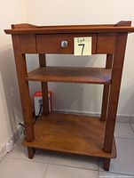 Photo of a medium brown wooden stand with one drawer and two shelves, set against a white wall and tiled floor.