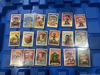 Image showing 18 Garbage Pail Kids trading cards from 1985-1987 in protective sleeves laid out in rows on a blue background.