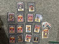 Photo showing multiple Garbage Pail Kids cards laid out on carpet, various characters visible, cards in protective sleeves and loose.