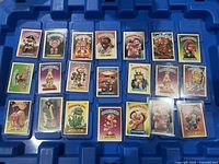 Top-down view of 21 Garbage Pail Kids cards placed on a blue tray, each in protective sleeves, showing colorful cartoon characters with the Garbage Pail Kids branding and character names.