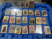 Overview of all 25 Garbage Pail Kids cards arranged on blue tray, showing front-facing colorful illustrated collectible trading cards from 1985-1987, each in protective sleeves.