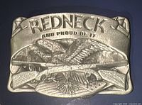 Front view of metal belt buckle showing "REDNECK AND PROUD OF IT" text and design of two ducks and crossed rifles.
