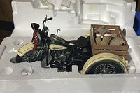 Side view of the 1:10 scale 1947 Harley-Davidson Servi-Car model in styrofoam packaging showing the green and cream paint, black seat, front fender with red bow, and a wooden crate at the back with delivery sacks.