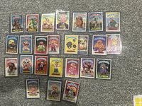 Top-down photo showing all 25 Garbage Pail Kids cards fanned in rows on a carpeted surface. Each card is in a clear plastic protective sleeve. Cards display various colorful cartoon characters with humorous names.