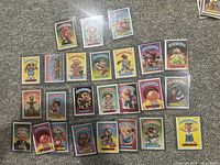 25 Garbage Pail Kids cards laid out on carpet in two images showing front of each card in plastic sleeves.