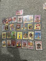 Lot of 30 colorful Garbage Pail Kids trading cards displayed on carpet, each sleeved in plastic. Various characters with different cartoonish styles and humor.