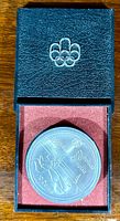 Silver coin with sailing motif visible inside plastic capsule placed within black textured case showing Olympic rings emblem.